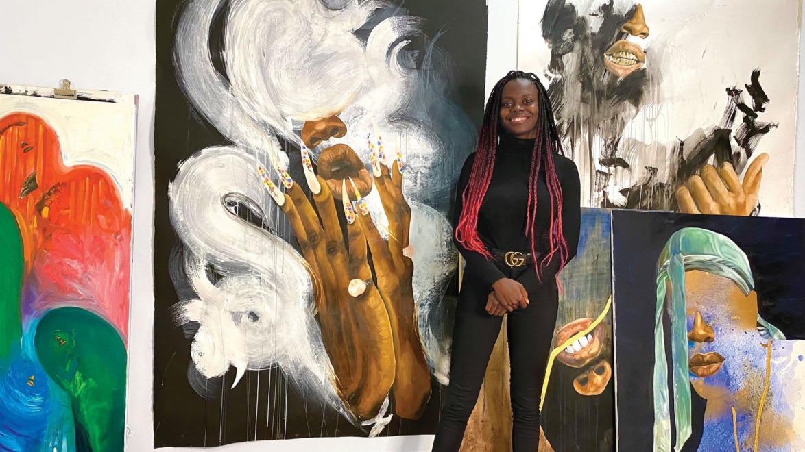 Artist standing in front of colorful, expressive paintings displayed on a white wall.