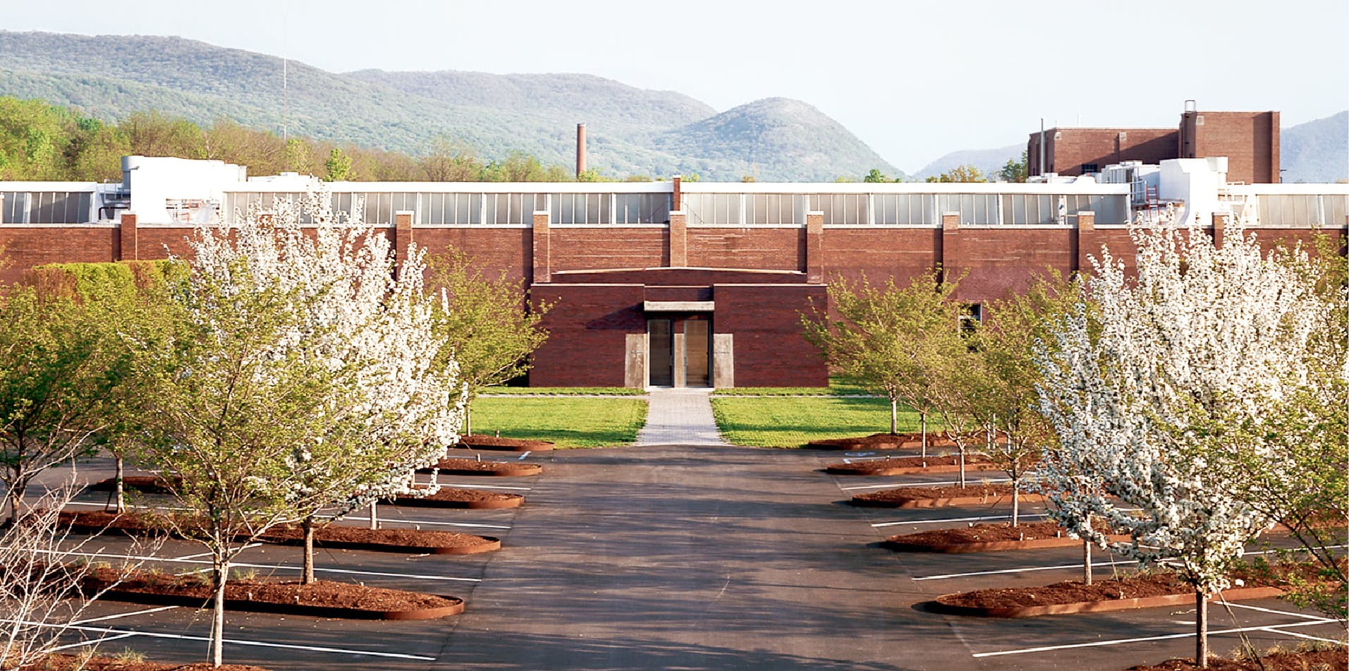 Modern industrial building surrounded by blooming trees and mountains in the background on a clear day.