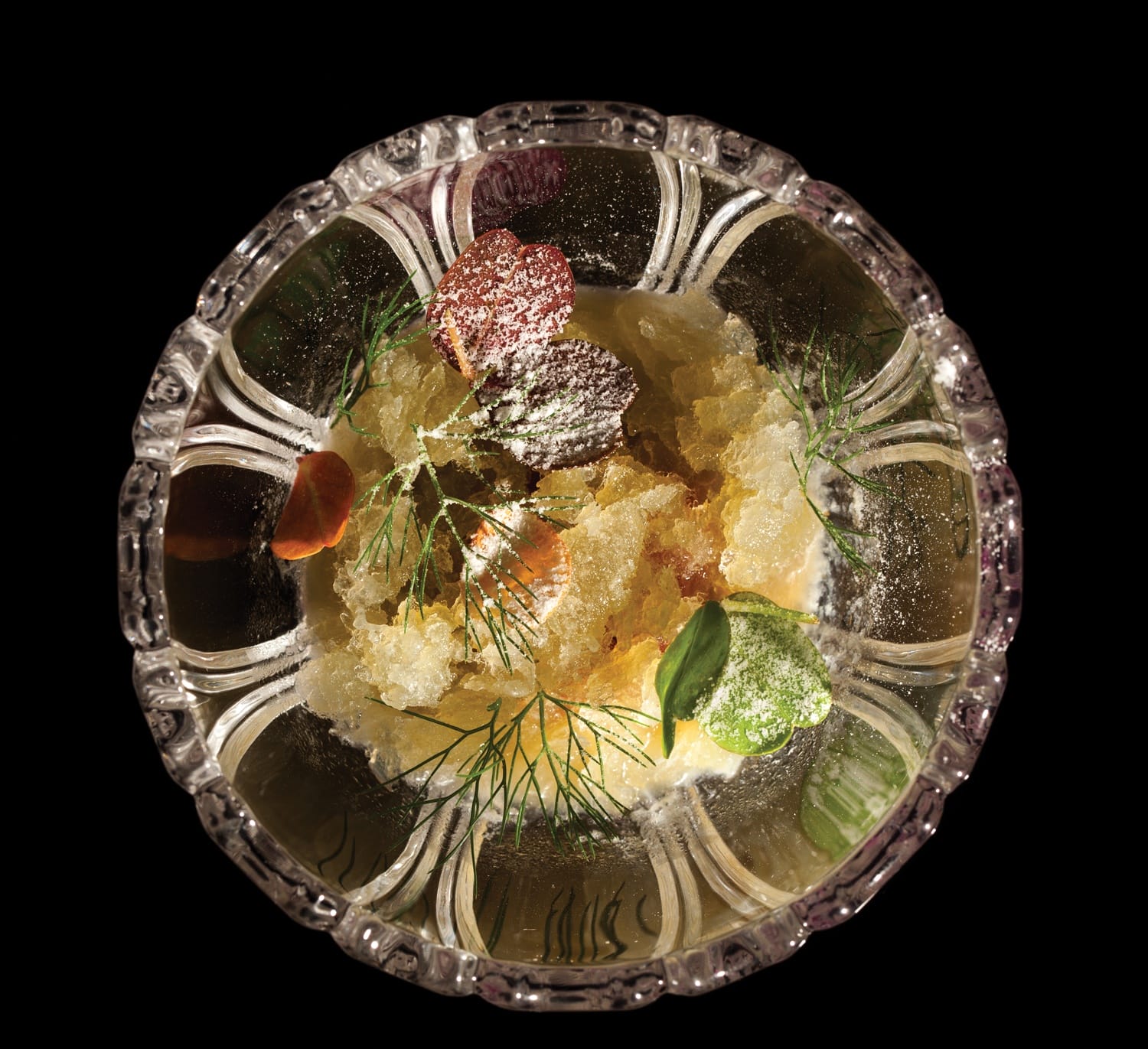 Crystal bowl with powdered leaves, herbs, and a translucent substance on a black background.