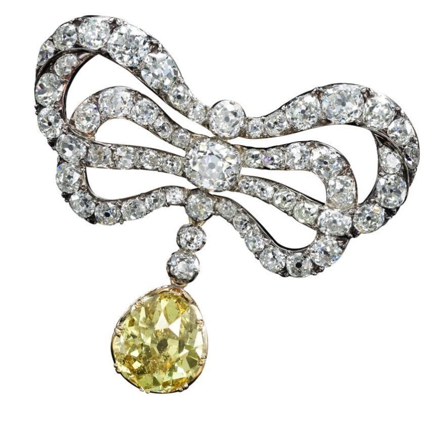 Antique diamond bow brooch with a large yellow gemstone pendant hanging in the center against a plain white background.