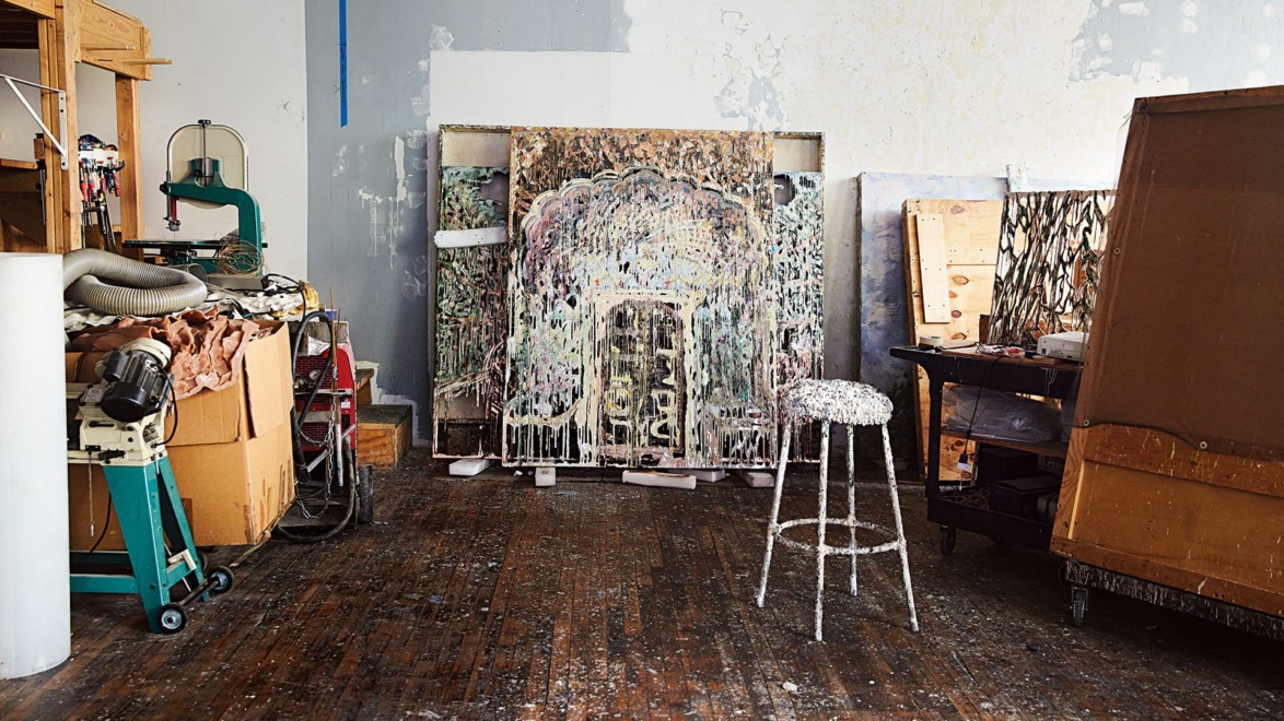Artist's studio with paint-splattered stool, canvases, tools, and materials scattered on a wooden floor.