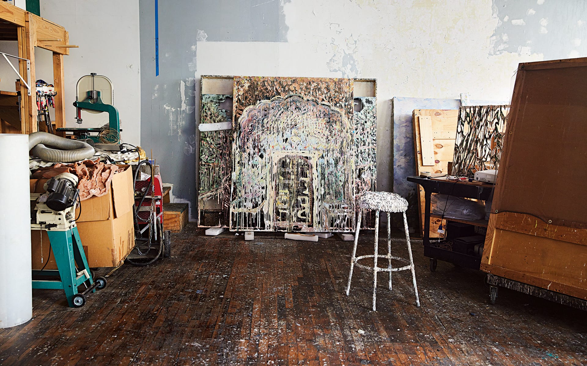 Artist's studio with paint-splattered stool, canvases, tools, and materials scattered on a wooden floor.