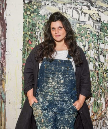 Person in paint-splattered overalls stands in front of an abstract, colorful painting on a wall.