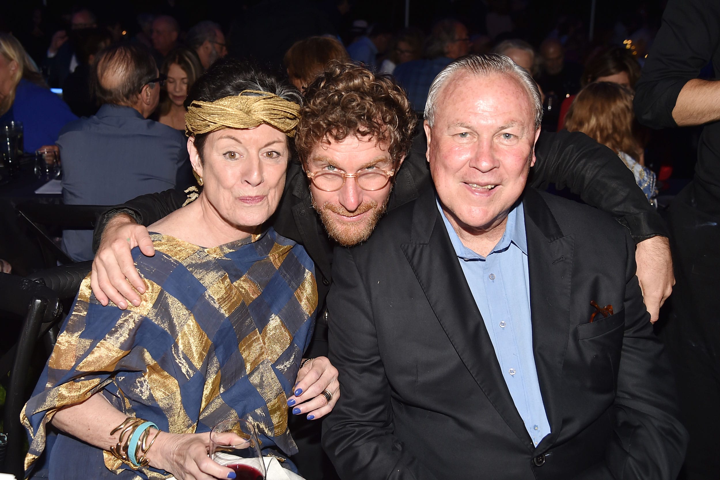 Three people smiling at a party, seated closely together with others in the background.