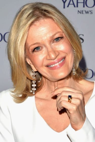 Smiling woman with shoulder-length blonde hair, wearing dangling earrings and a blue stone ring, at a news event backdrop.