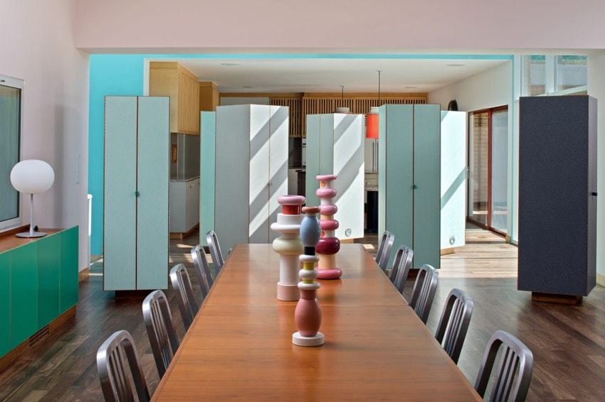 Modern dining room with a long wooden table, colorful vases, metal chairs, and tall cabinets in the background.