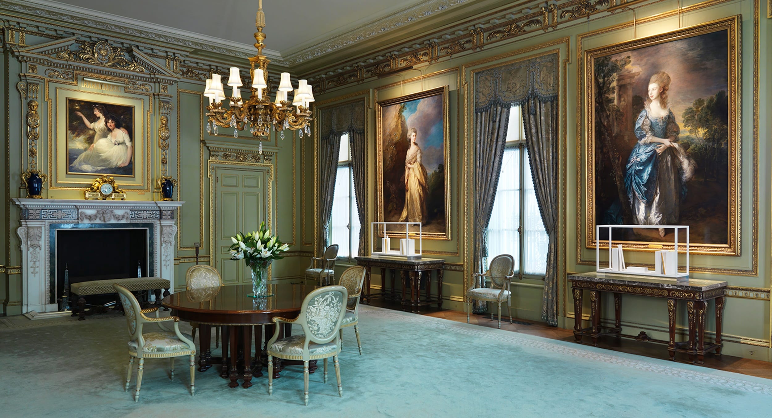 Elegant room with ornate decor, chandelier, and large paintings on walls, featuring classical furniture and a decorated fireplace.