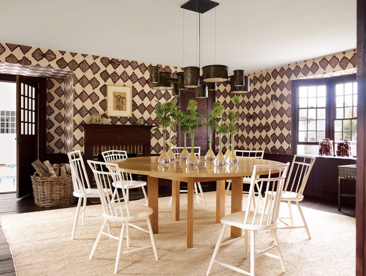 Modern dining room with wooden table, white chairs, geometric wallpaper, and decorative plants in vases on the table.