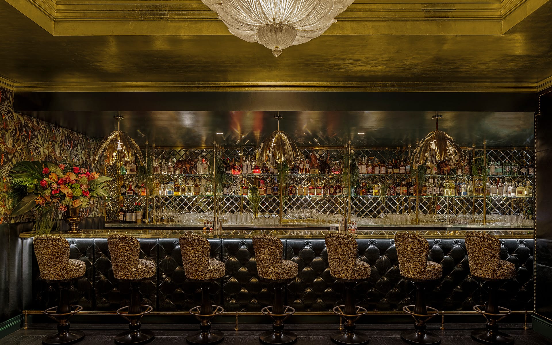 Luxurious bar interior with patterned bar stools, glass chandeliers, and an elegant floral backdrop behind a stocked counter.