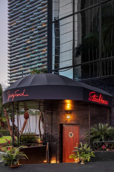 Entrance to a modern steakhouse with a black awning and neon red sign, surrounded by palm trees and glass building reflections.