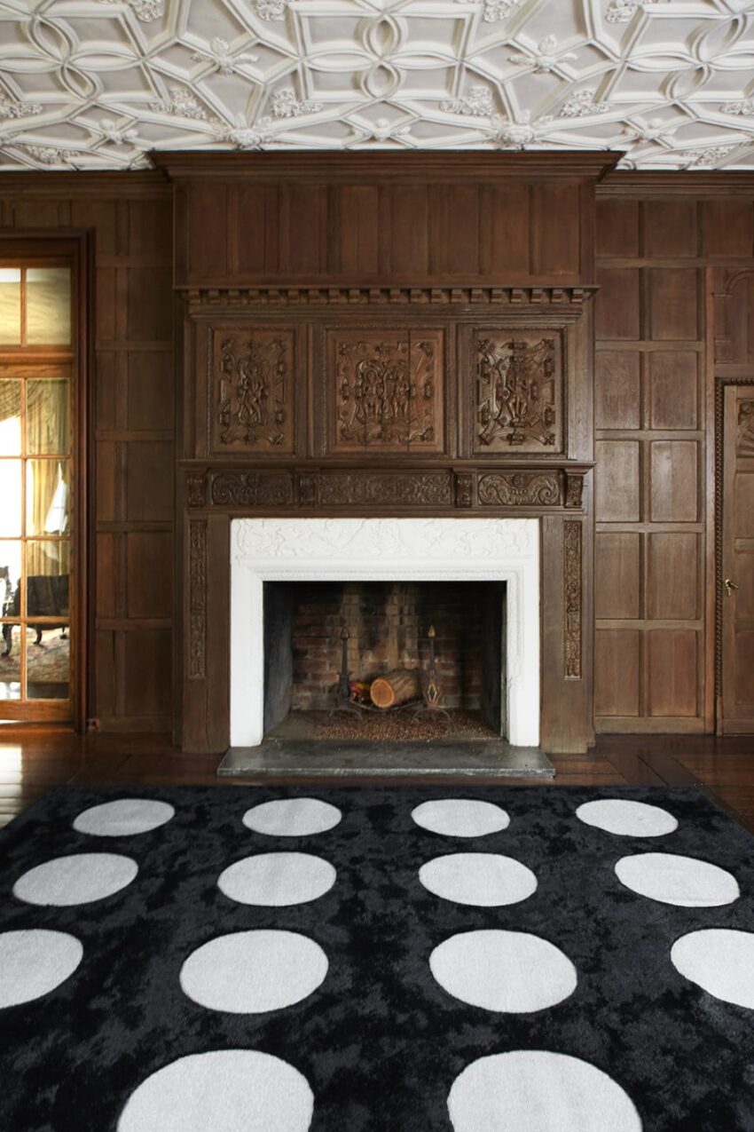 Ornate wooden fireplace with intricate carvings, white mantel, black rug with large white circles, and detailed ceiling design.