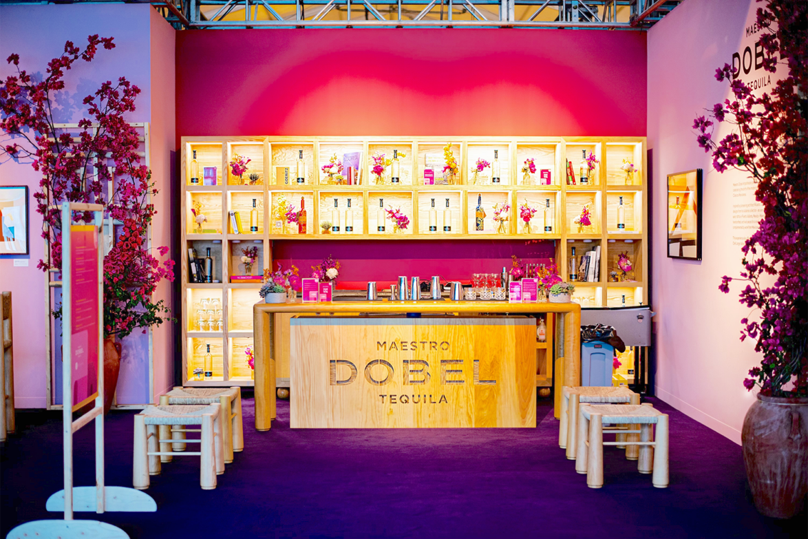 Bar setup with pink and purple theme, wooden counter, stools, and illuminated shelves displaying tequila bottles and pink flowers.