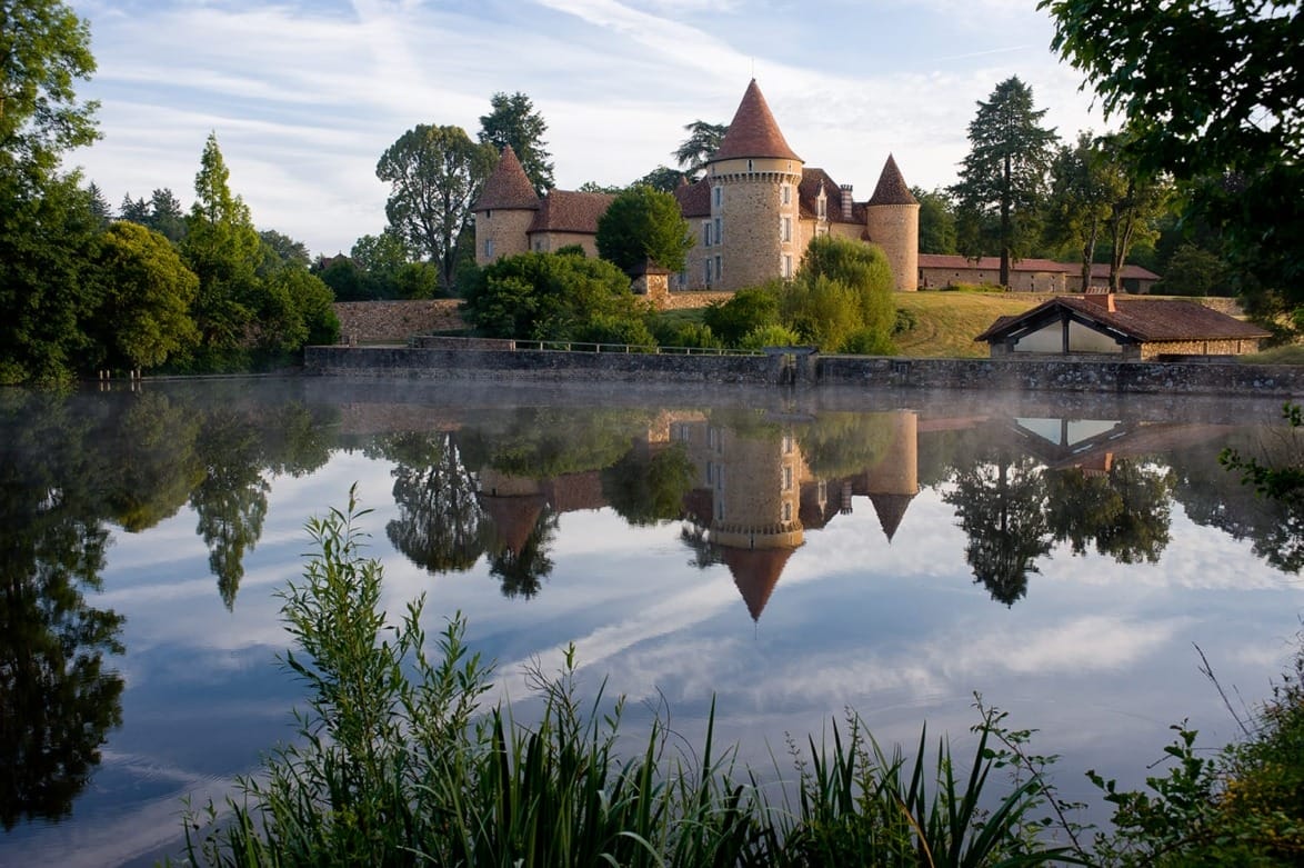 Castle with round towers and spires reflected in a calm lake, surrounded by trees and greenery under a blue sky.