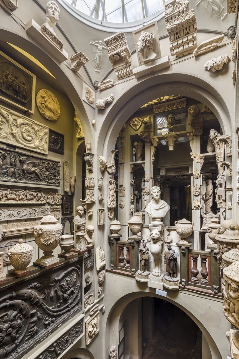 Museum interior with classical sculptures, busts, reliefs, and architectural elements displayed on multi-level arches.