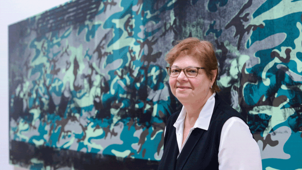 Person smiling in front of a large abstract artwork with blue and green camouflage patterns in a gallery setting.