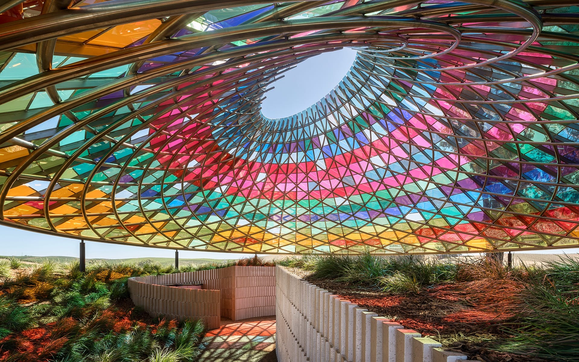 Colorful glass structure with spiral design casting vibrant shadows on surrounding landscape and pathway.