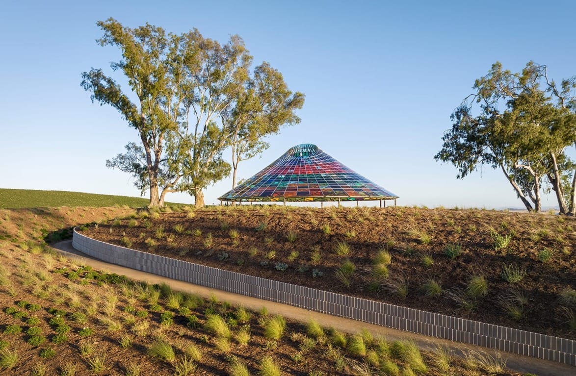 Colorful glass dome structure surrounded by trees and plants on a sunny hillside landscape.