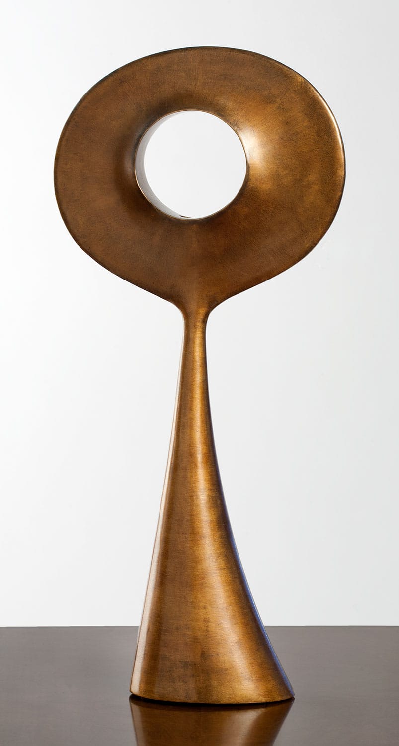 Abstract bronze sculpture with an elongated base and circular top featuring a central hole, displayed on a flat surface.