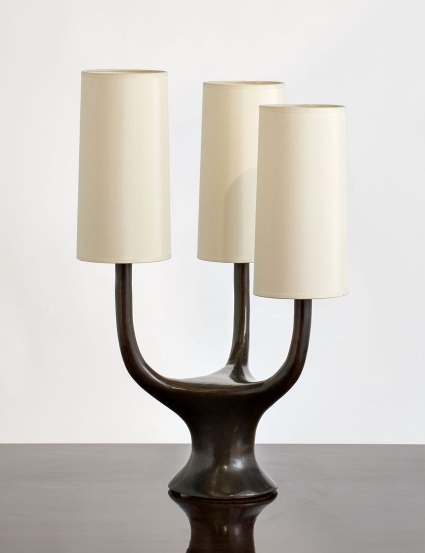 Three-light bronze candelabra table lamp with cream drum shades on a wooden surface.