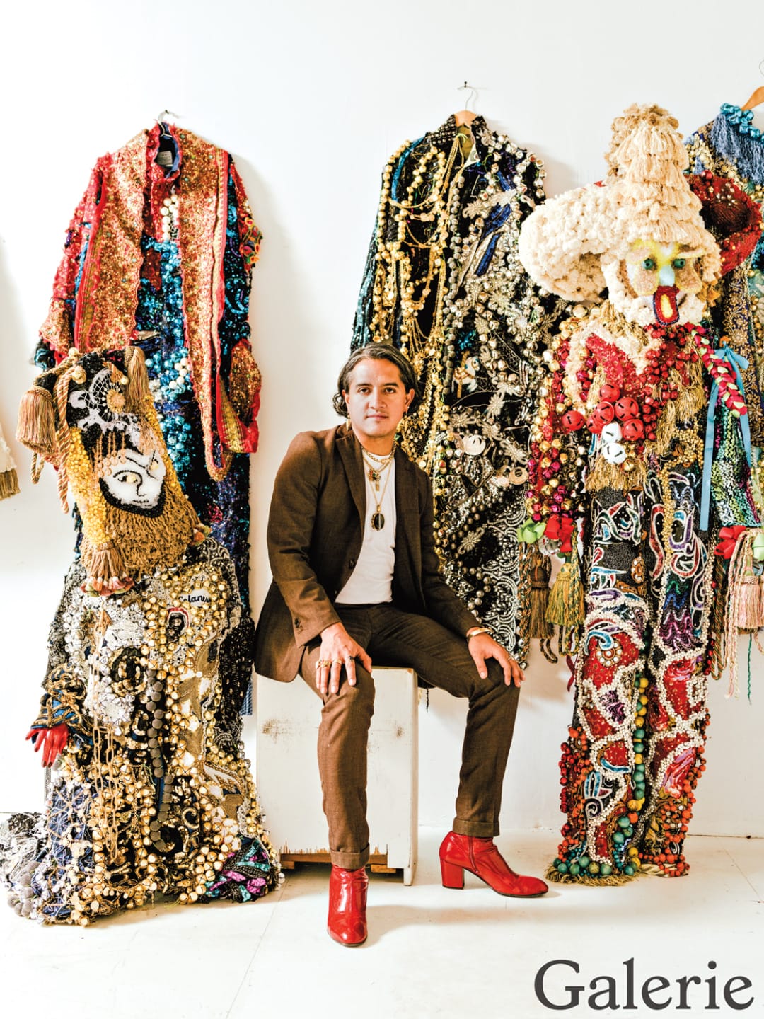 Person seated among vibrant, intricately designed costumes with beaded and textured embellishments on display in a gallery setting.