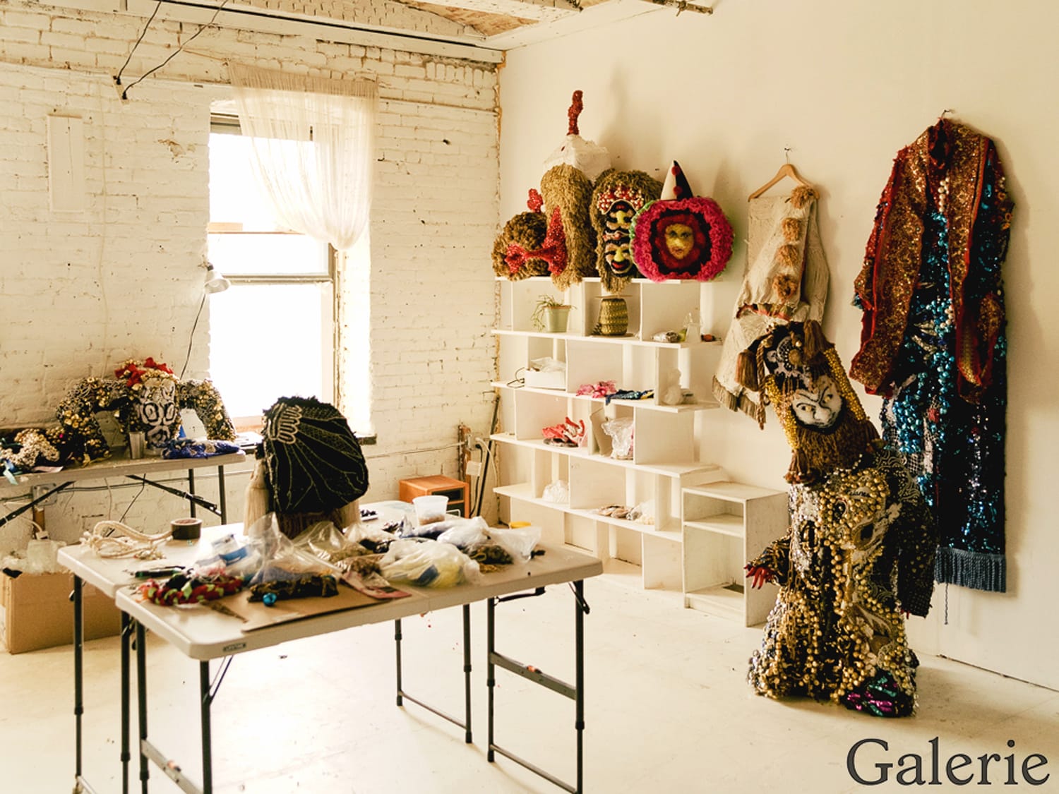 Art studio with elaborate masks and costumes on shelves and hanging, table with crafting materials, window light streaming in.