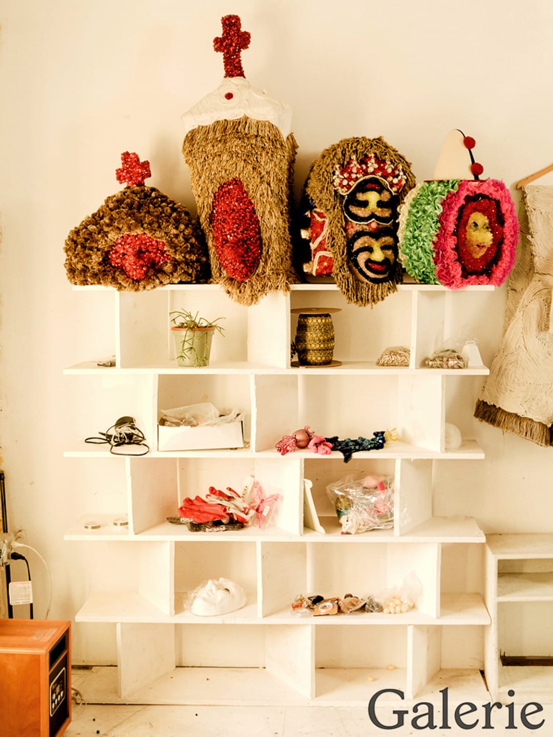 Colorful handmade masks displayed on white shelves with various small crafts and decorations nearby in a gallery setting.
