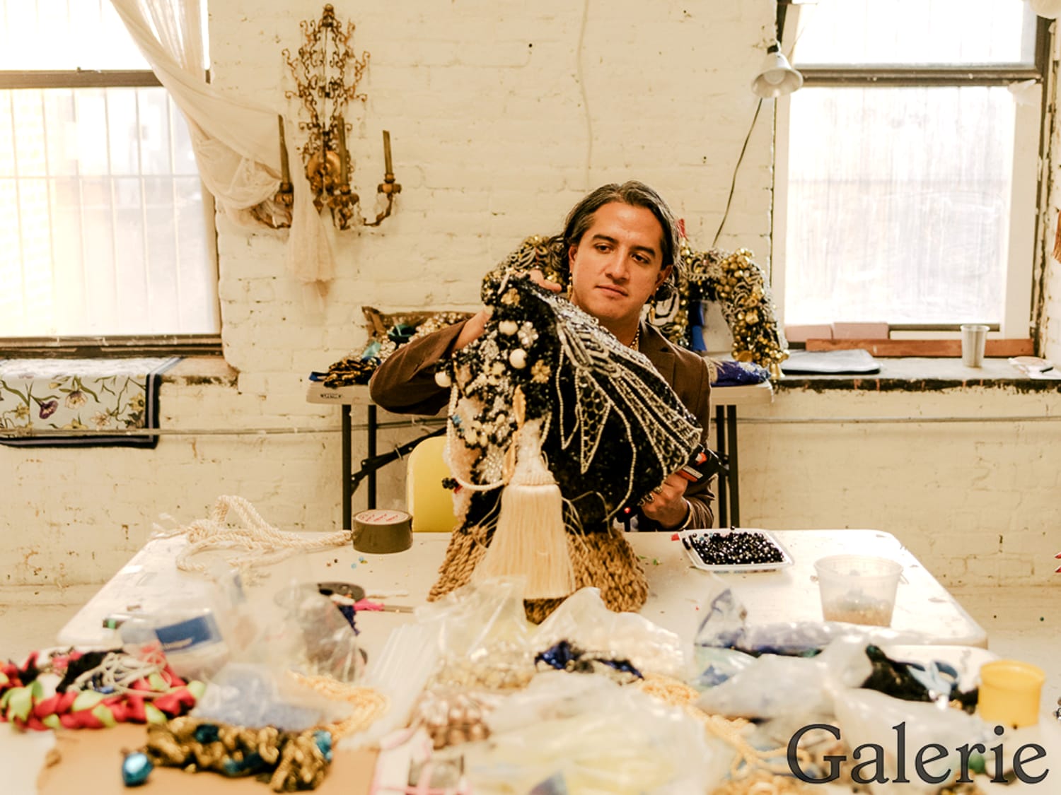 Person working with beads and fabric at a craft table in a sunlit room with large windows.