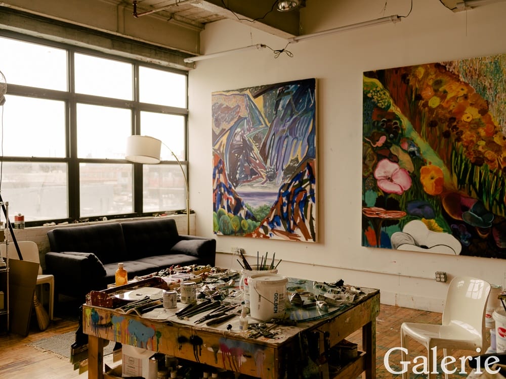 Artist studio with colorful paintings on the walls, a workbench with art supplies, and a large window letting in natural light