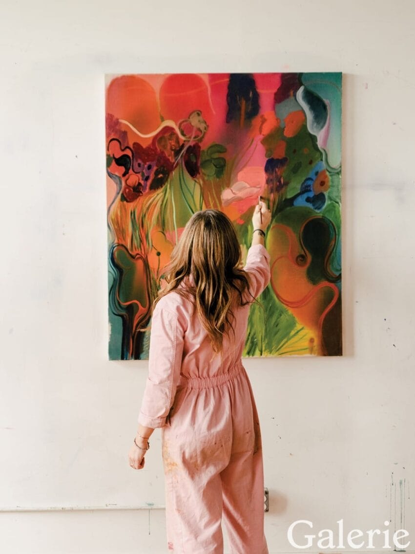 Artist in pink overalls painting a colorful abstract artwork on a large canvas in a studio setting.