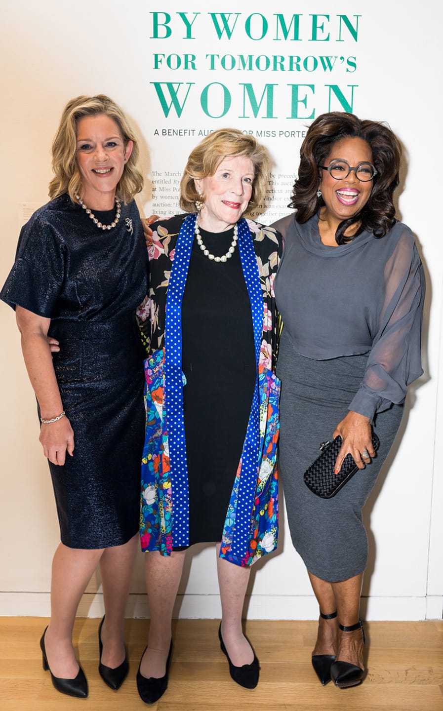 Three women standing together, smiling, in front of a sign that reads "By Women For Tomorrow's Women" at a benefit event.