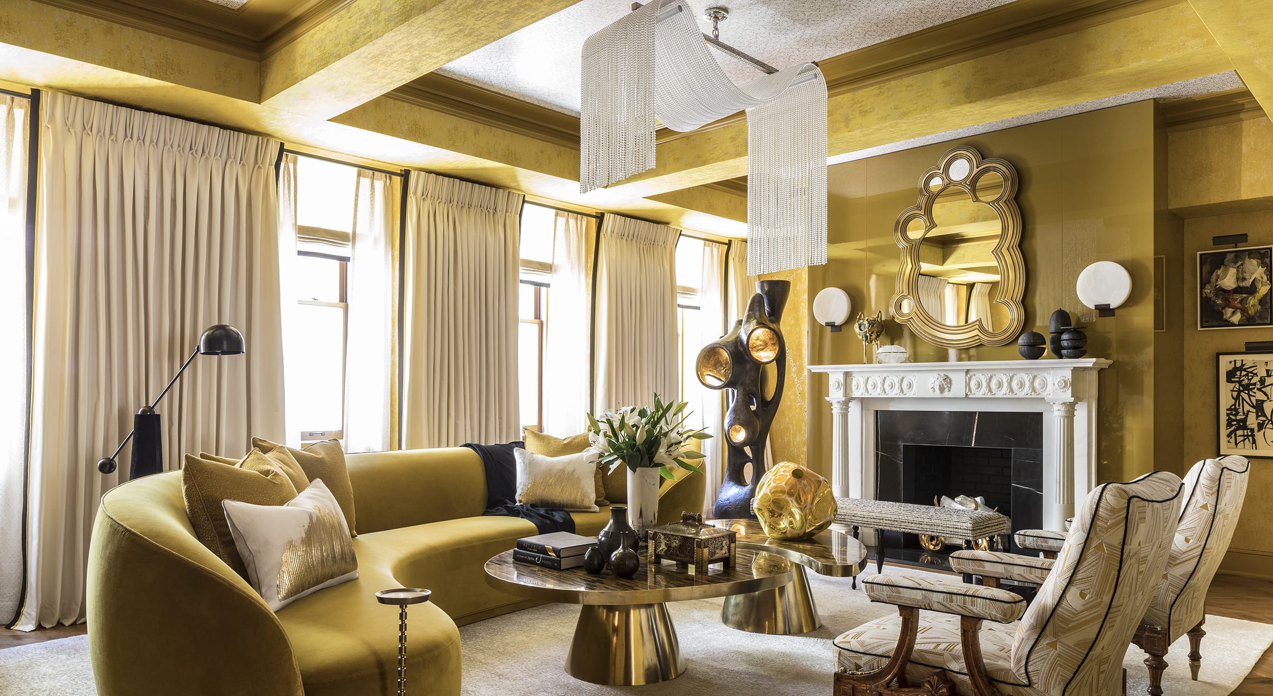 Elegant living room with gold accents, curved sofa, modern chandelier, artistic decor, and a fireplace with a unique mirror above.
