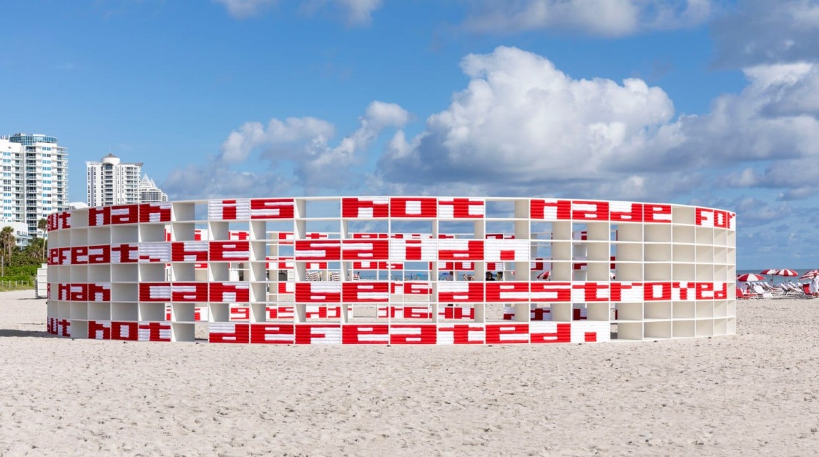 Art installation on a sandy beach with a circular structure displaying red text and tall buildings in the background.