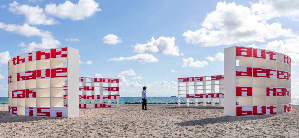Art installation on a sandy beach with red and white text structures against a blue sky background, person observing.