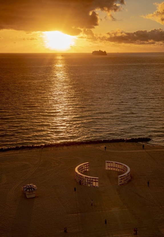 Sunset over ocean with a distant ship, sandy beach in foreground featuring a circular art structure and people walking.