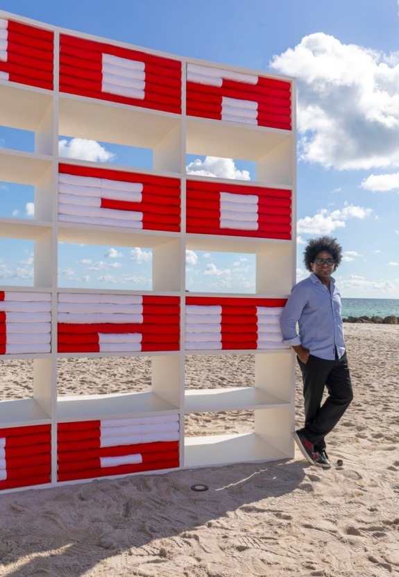 Person standing beside a shelf of neatly stacked red and white towels on a sunny beach.
