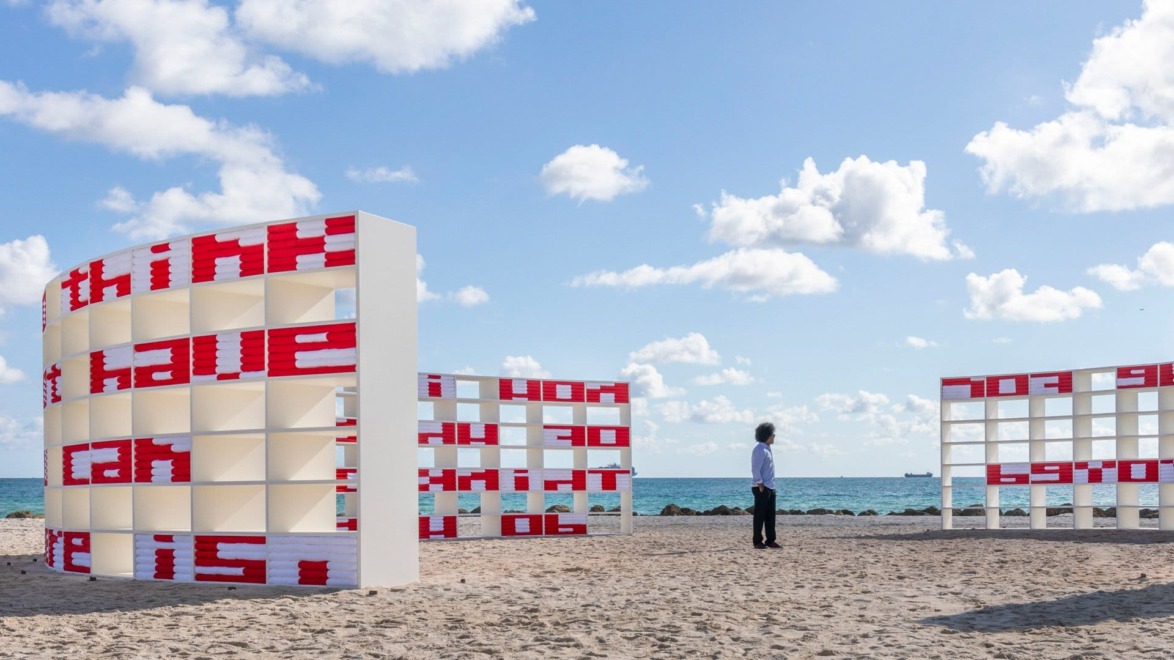 Art installation with curved structures and red text on a sandy beach, blue sky with clouds in the background.