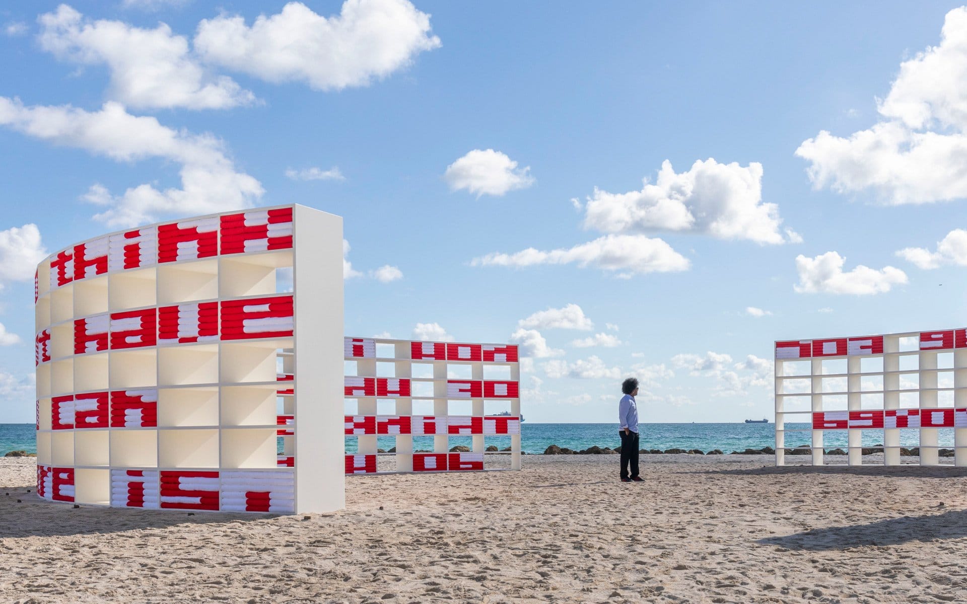 Art installation with curved structures and red text on a sandy beach, blue sky with clouds in the background.