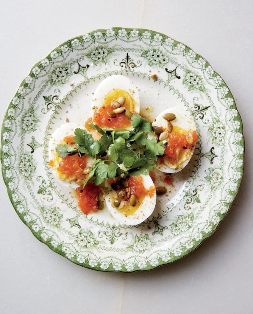 Decorative plate with sliced boiled eggs topped with tomato salsa, cilantro, and seeds on a white background.