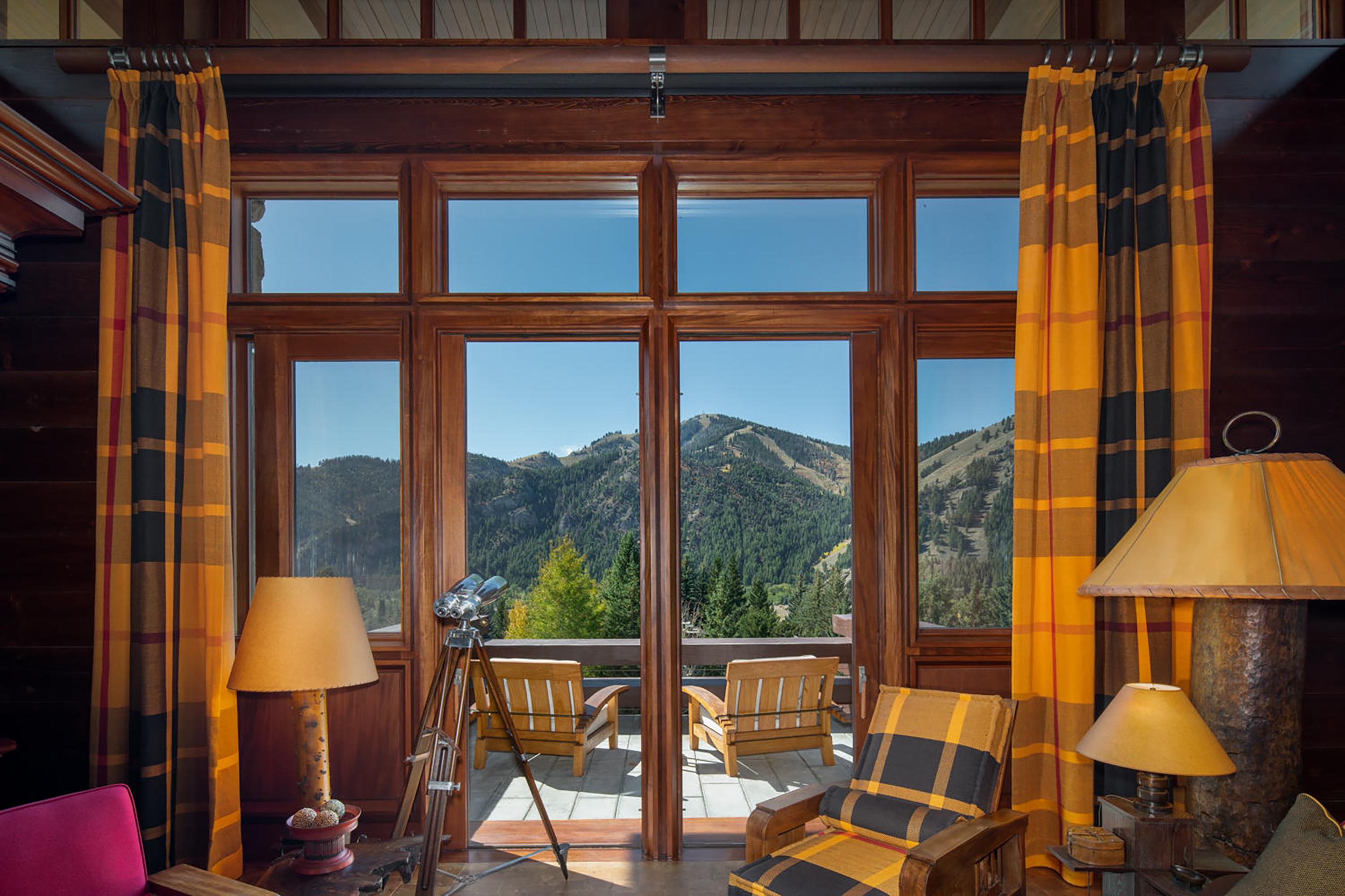 Interior view with wooden framed windows, yellow plaid curtains, telescope, chairs outside, and mountain scenery in the background.