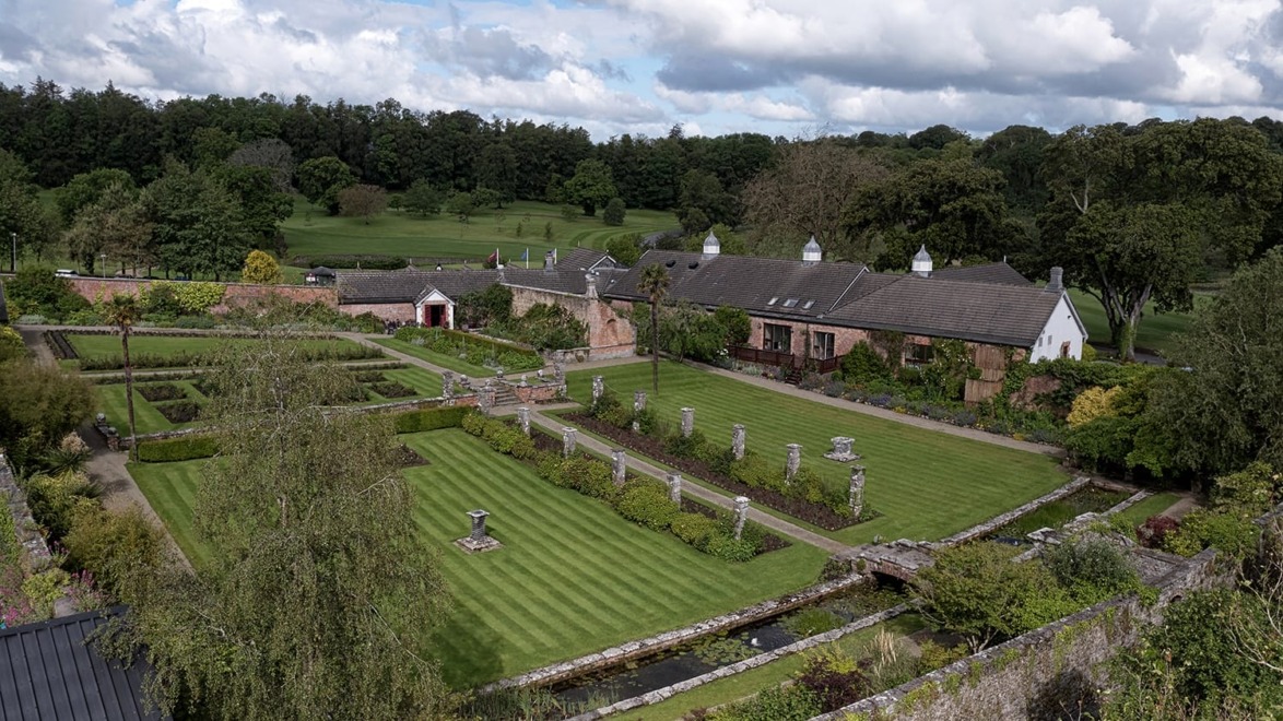 Aerial view of a garden with manicured lawns, stone pathways, hedges, and a brick building in the background.