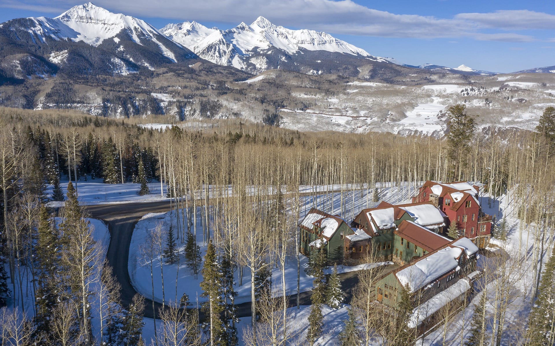 Scenic snowy mountain landscape with rustic buildings, tall trees, and a winding road under a clear blue sky.
