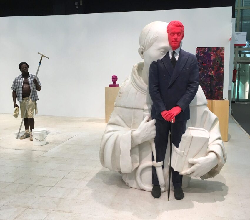 Modern art exhibit featuring a painted red figure in a suit, blending with a large white statue in an abstract setting.