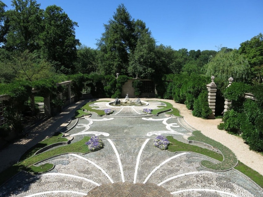 Elegant garden with intricate stone patterns, lush greenery, and a central fountain under a clear blue sky.