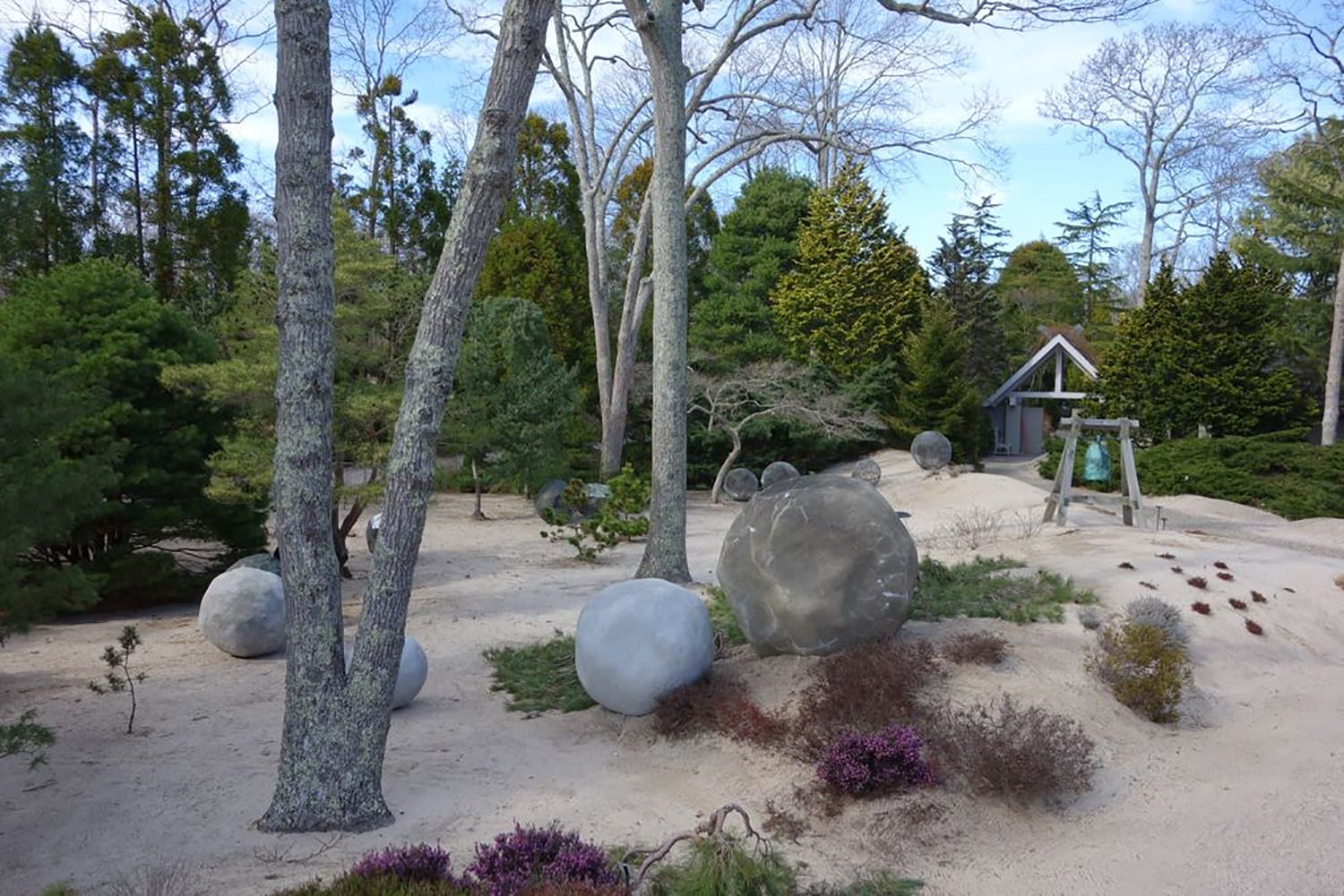Zen garden with large stone spheres, trees, shrubs, sand, and a small structure in the background.