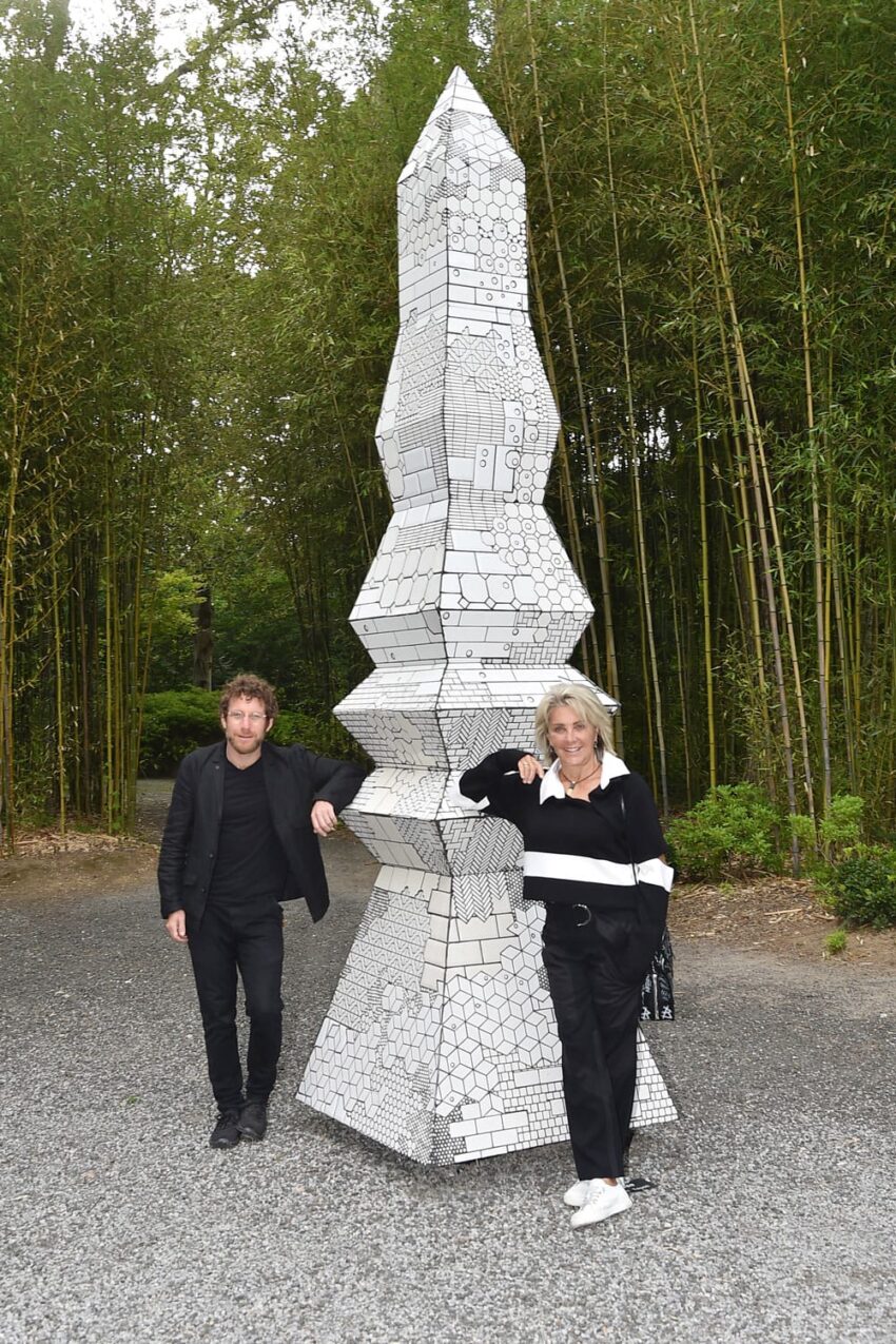 Two people posing beside a tall geometric sculpture in an outdoor setting with bamboo in the background.