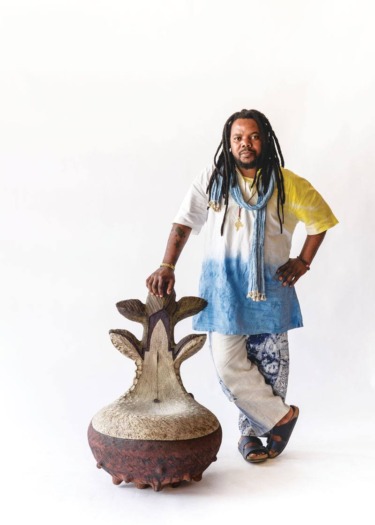 Man in colorful tunic and necklace stands beside a unique chair with intricate design against a white background.