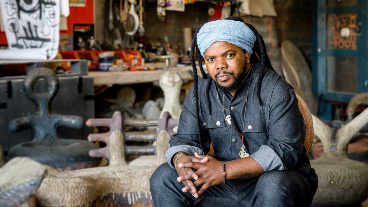Artist seated in a studio surrounded by sculptures and art supplies, wearing a blue headwrap and denim outfit.