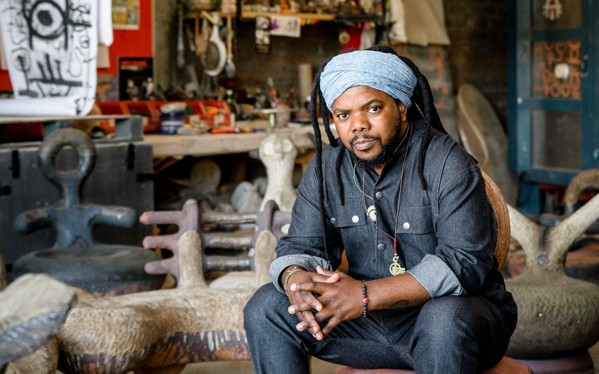 Artist seated in a studio surrounded by sculptures and art supplies, wearing a blue headwrap and denim outfit.
