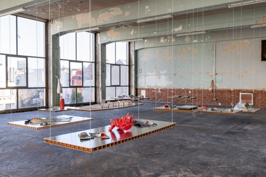 Industrial art installation with suspended tables, coral sculpture, and views of urban buildings through large windows.