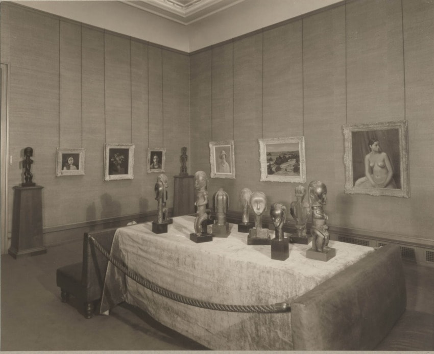 Art gallery with sculptures on a covered table and framed paintings on the walls in a softly lit room.